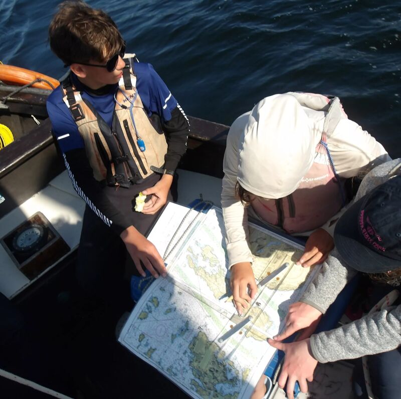 The image shows three people on a boat looking at a map. One person is standing and wearing a life vest and sunglasses, while the other two are seated and pointing at the map. The boat is on the water, and the overall scene suggests they are navigating or planning a route. The map appears to be a nautical chart, possibly for sailing or boating.